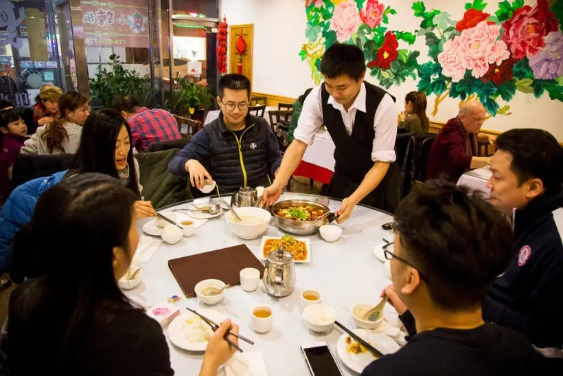 一群人在装饰有花卉壁画的餐厅内用餐，圆形餐桌上摆满餐具和美食，六人围坐享受，左侧黑衣男士与对面女士专注看手机，中央服务员正上菜，背景是其他顾客和餐厅装饰，营造忙碌而温馨氛围。