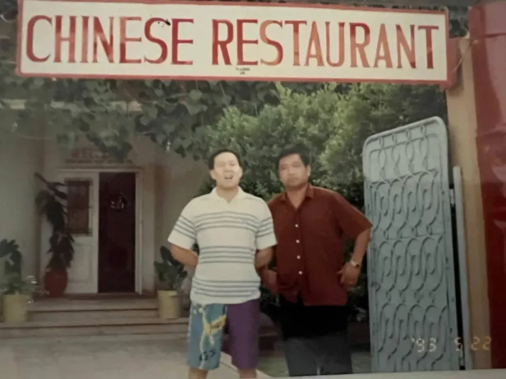 两位男士微笑着站在一家名为“CHINESE RESTAURANT”的餐厅前合影，背景中有半开的门、植物以及餐厅横幅和日期“93年8月”，营造出怀旧氛围。