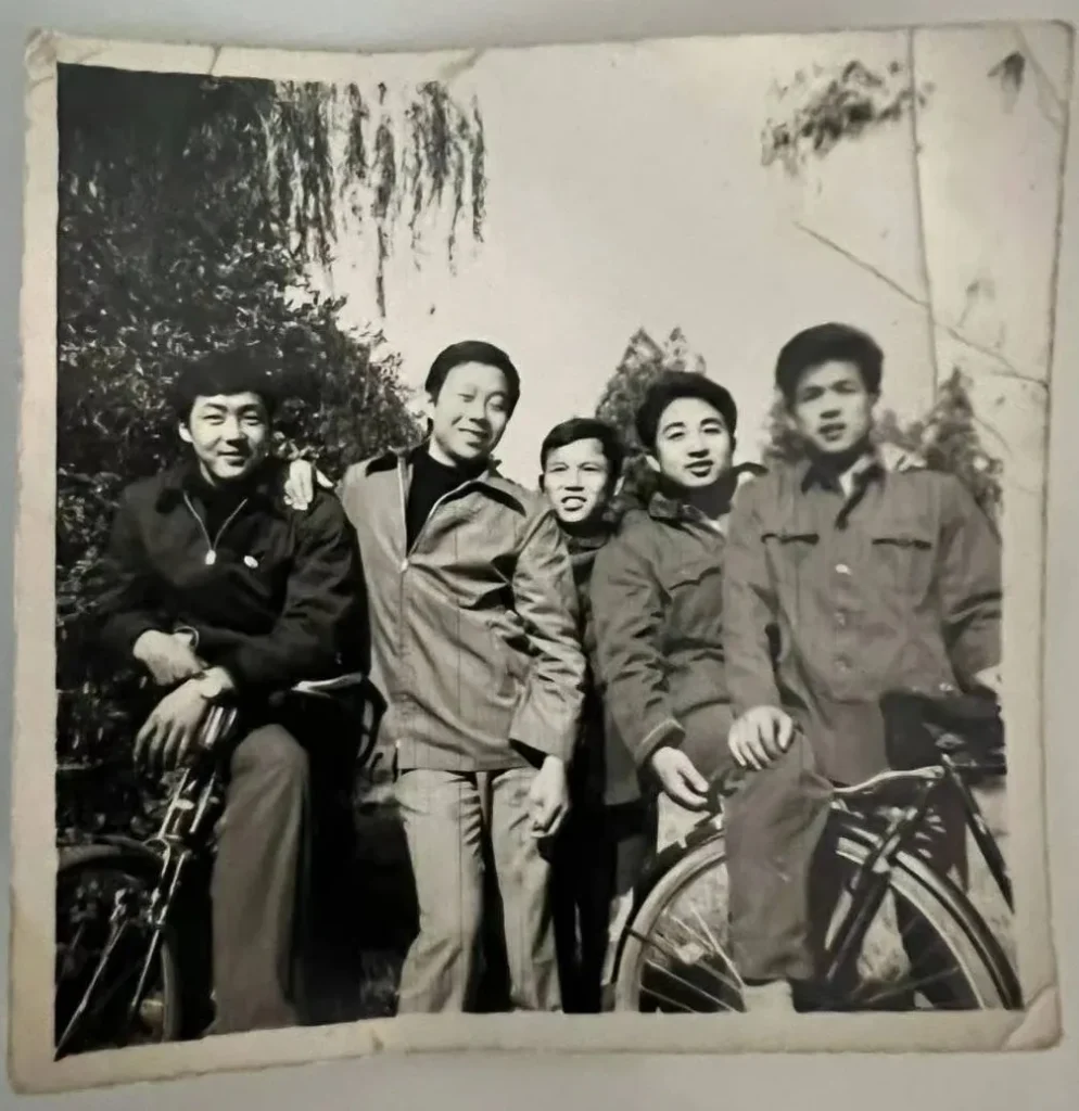 黑白老照片中，五位亚洲男性在户外合影留念。他们穿着深色衣物，从左至右，前四位男士分别微笑看向镜头、微笑、表情严肃、面带微笑扶自行车，最后一位男士站在自行车旁微笑。背景为模糊的树木和灌木丛，增添历史感。
