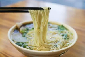 一碗热气腾腾的兰州牛肉面，面条被筷子夹起，清汤中漂浮着葱花和蔬菜，展现了手拉面的细致工艺。