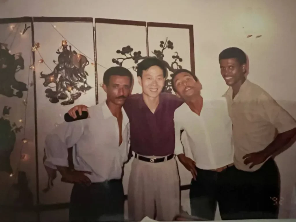 四位男士在室内合影，站在带有花卉图案及“大吉”字样屏风前，背景为白色墙壁和装饰品。他们分别穿着白色、紫色、浅色和黑色短袖衬衫，搭配深色裤子，面带微笑，表情自然，姿态轻松，营造出温馨友好的氛围。