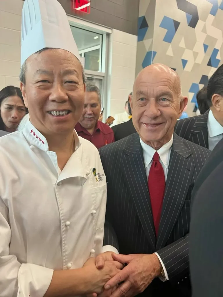 两位男士在室内场合握手，左边是穿着厨师服并戴着厨师帽的厨师，面带微笑，右边则是穿着西装打着领带的男士，同样面带微笑。他们背后有其他人，但面部表情不清晰。背景装饰有蓝色和白色的几何图案，整个场景正式而友好。