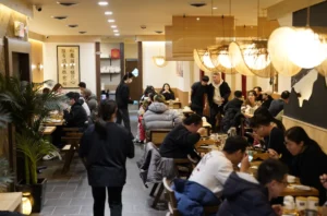 "这张照片展示了一个装饰简约而现代的餐厅内部，以木质元素为主，墙上挂有中国书法作品'腹有詩書技愈精'（部分文字，可能因分辨率限制无法完整辨认）。餐厅内，顾客们坐在长方形木桌旁，有的在用餐，有的在交谈，氛围热闹而温馨。桌上摆有餐具和饮料，墙上还装饰有灯笼和植物，营造出一种舒适的用餐环境。"