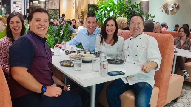 一群人在餐厅愉快用餐，桌上摆满美食与饮料，手机置于桌上，记录这美好时光，背景绿意盎然，增添用餐氛围。