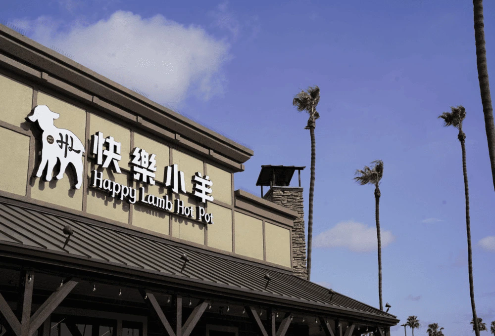 蓝天白云下，一栋白灰色建筑立面，醒目的“快乐小羊 Happy Lamb Hot Pot”门头