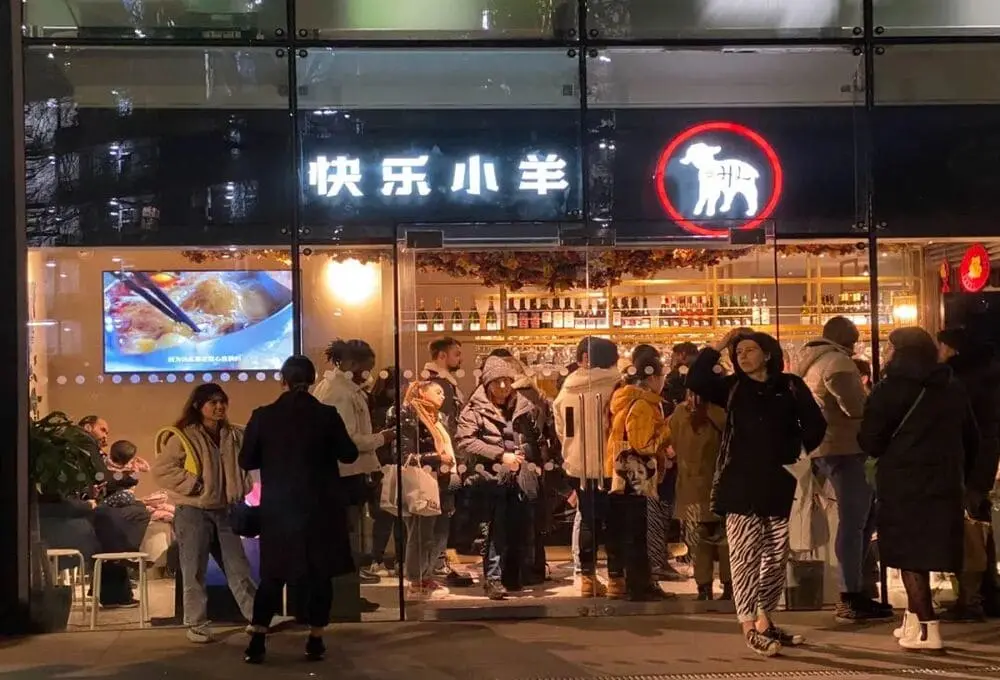 夜幕下的街道上，一栋玻璃建筑立面，醒目的“快乐小羊 ”门头灯箱，一群不同族裔在排队的等待就餐的食客