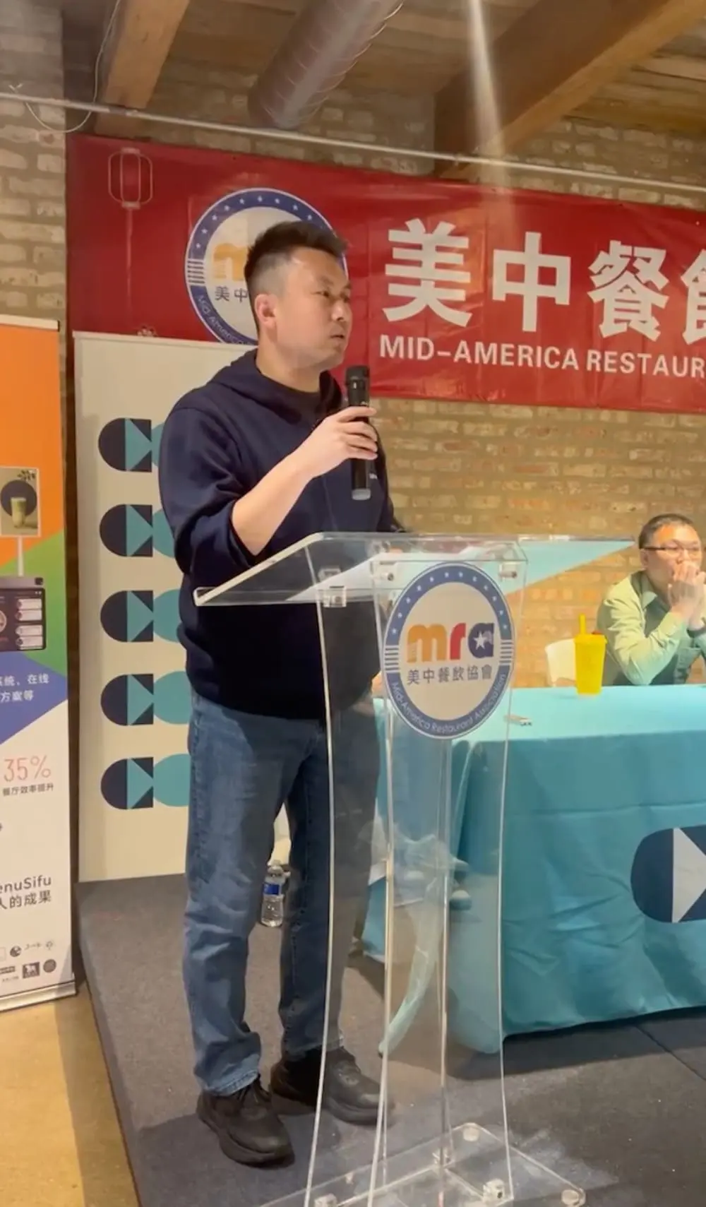 身着深色卫衣的Alex Ma手持麦克风，在透明讲台前发表演讲，背景可见“美中餐饮协会”横幅和MenuSifu宣传海报。