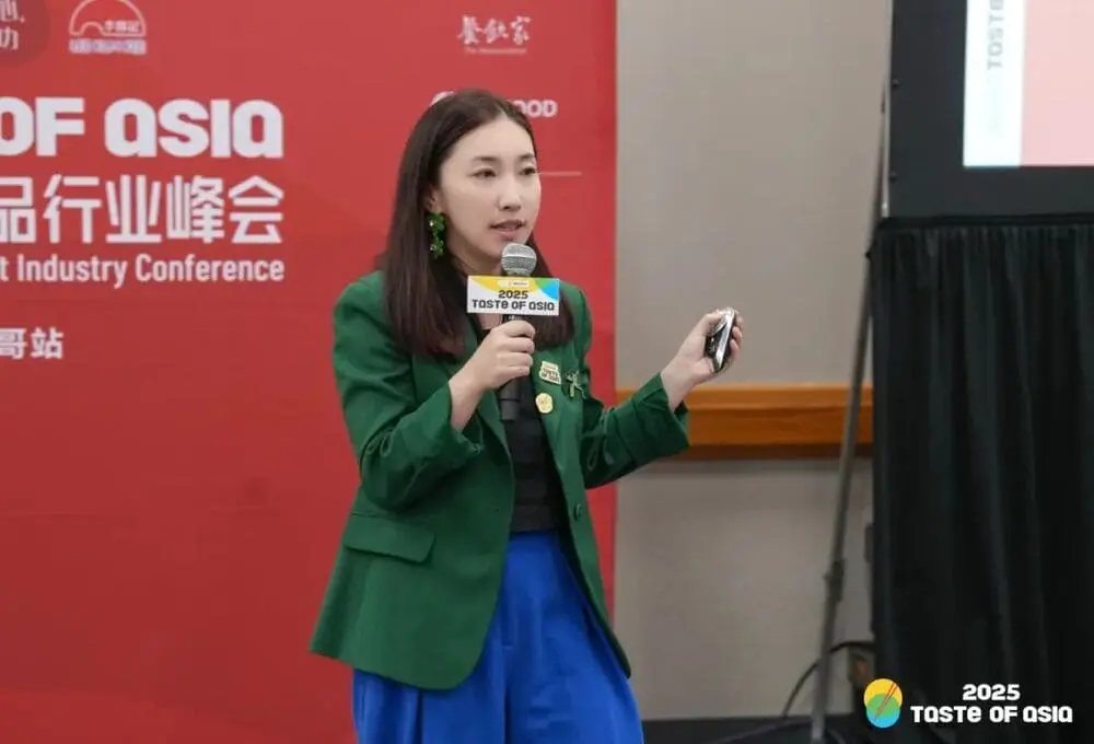 身穿绿色西装的演讲嘉宾手持麦克风，在“2025 Taste of Asia”峰会芝加哥站演讲，背景为带有会议标识的红色展板，场地为会议厅。