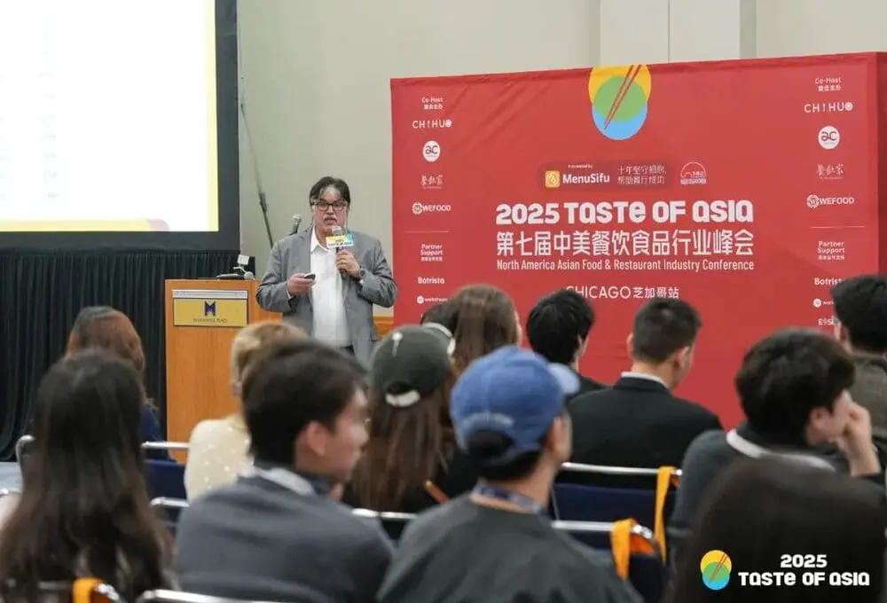 演讲嘉宾手持麦克风，在“2025 Taste of Asia”峰会芝加哥站演讲，背景为带有会议标识的红色展板，场地为会议厅。嘉宾面对前面很多观众认真听讲