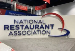 美国全国餐馆协会（NRA）展会品牌标识墙：白底蓝字立体呈现"NATIONAL RESTAURANT ASSOCIATION"，右侧红蓝弧形设计呼应品牌色，顶部红色横幅标注"WORKFORCE DEVELOPMENT"与"RESEARCH & INSIGHTS"，左侧"Serving Every"标语传递行业包容性，灰毯地面与高对比视觉凸显餐饮行业权威性与专业战略导向。