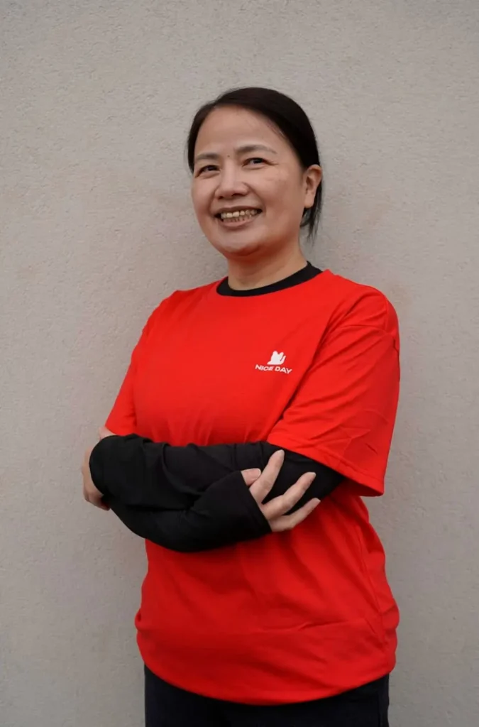 一位中年福建妈妈Joyce，面带微笑，双臂交叉，身穿红色 “Nice Day” T恤，背景为灰色墙面。
