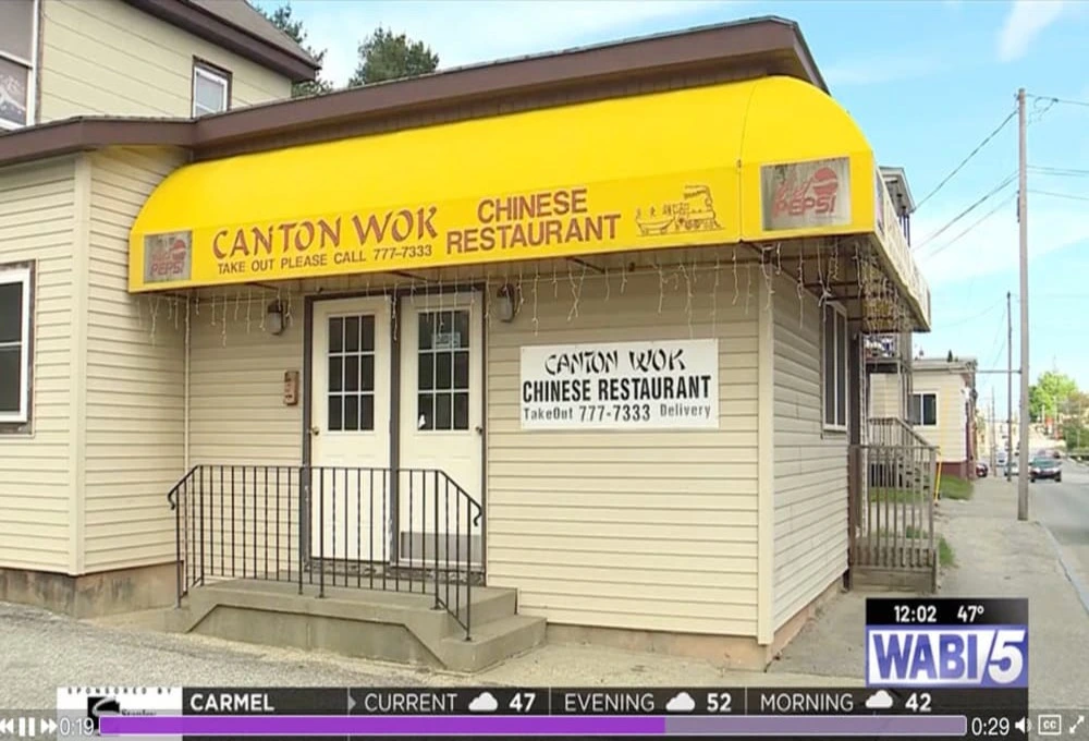 突关停的中餐馆外景：广式锅品（Canton Wok）位于缅因州刘易斯顿市里斯本街的店面实拍，米黄色外墙与黄色遮阳篷上印有订餐电话777-7333及部分拼写误差标识（如“CANTON WORK”）。画面捕捉到晴朗天气下的街景，橱窗已张贴出租告示，暗示该店因业主被ICE拘押而永久停业，与正文中Google页面关闭信息相印证。