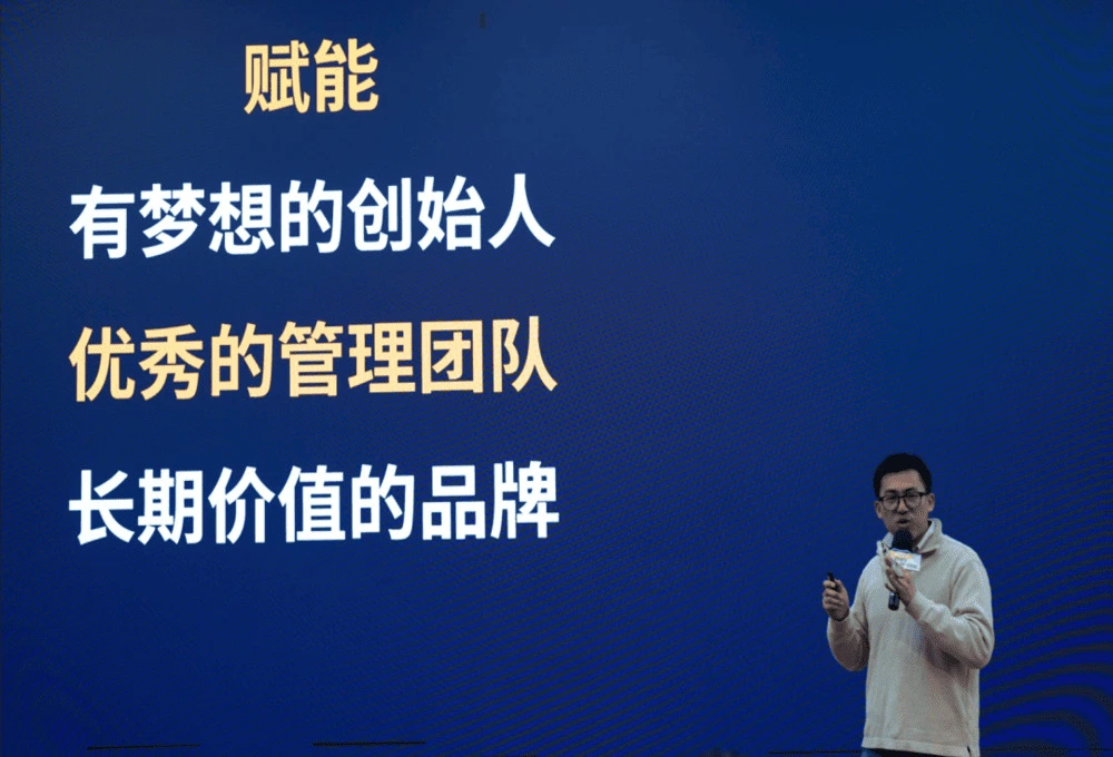 TOA峰会演讲现场：一位身着浅色衬衫的餐饮专家手持话筒，站在深蓝色背景前阐述企业成功要素。背景醒目展示三行白色文字"有梦想的创始人"、"优秀的管理团队"和"长期价值的品牌"，顶部黄色"赋能"二字点明主题。画面采用三分构图法，将演讲者与文字要素有机结合，背景渐变蓝色象征专业深度，黄色标题突出赋能理念，完整呈现餐饮企业从初创到规模化发展的核心人才与品牌建设要素，呼应文中MenuSifu提出的"六边形战士模型"中组织管理与品牌建设维度。