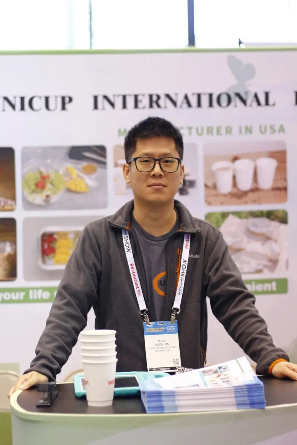 穿灰色外套的男士站在Nicup的广告版前，免签是一叠产品介绍彩页和一叠杯子样品