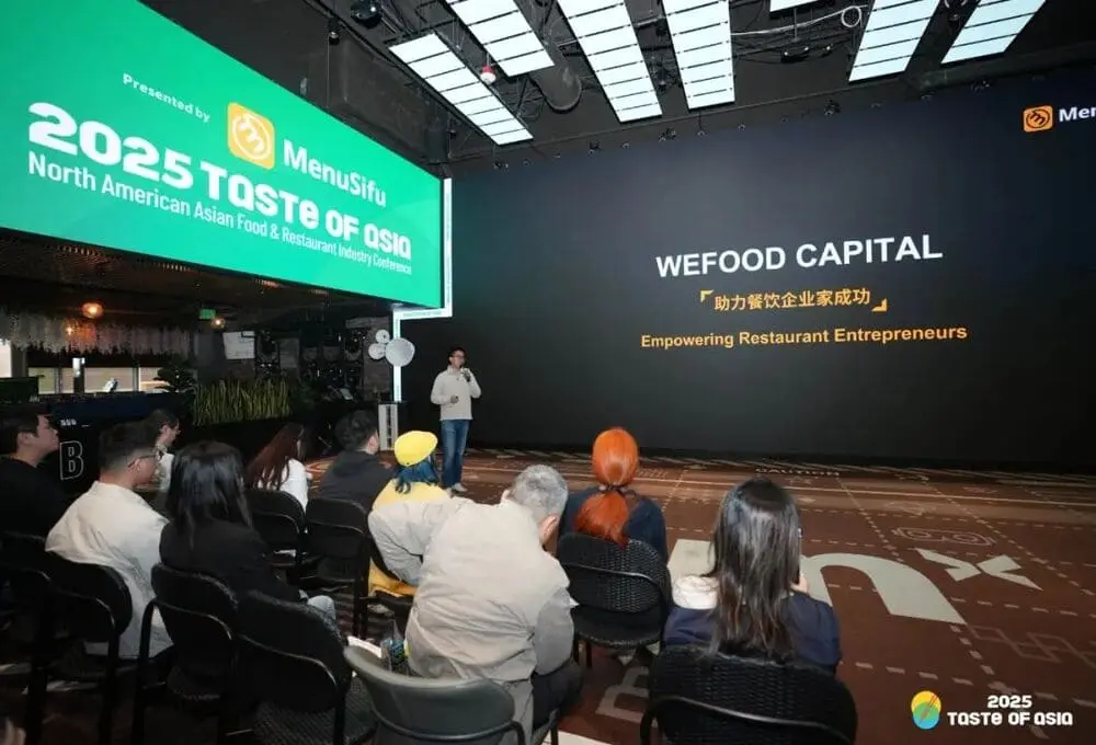 2025 TASTE OF ASIA峰会现场，MenuSifu高管在舞台中央发布WEFOOD CAPITAL餐饮基金。背景大屏幕清晰显示"WEFOOD CAPITAL"基金名称及"助力餐饮企业家成功/Empowering Restaurant Entrepreneurs"双语标语，左侧绿色横幅标注"2025 TASTE OF ASIA"活动名称及主办方MenuSifu标识。舞台采用现代几何地砖设计，深色墙面与矩阵顶灯营造专业氛围，台下坐满专注聆听的餐饮从业者。画面完整呈现北美首个专注中餐产业的5000万美元基金发布盛况，展现华人餐饮资本化进程的重要里程碑。