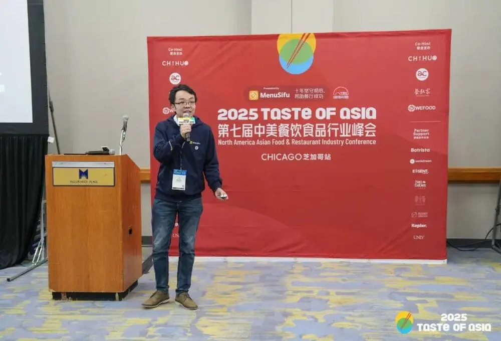 演讲嘉宾手持麦克风，在“2025 Taste of Asia”峰会芝加哥站演讲，背景为带有会议标识的红色展板，场地为会议厅。