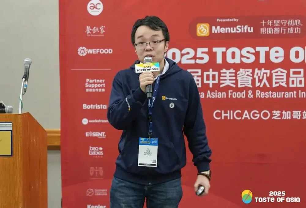 演讲嘉宾手持麦克风，在“2025 Taste of Asia”峰会芝加哥站演讲，背景为带有会议标识的红色展板，场地为会议厅。