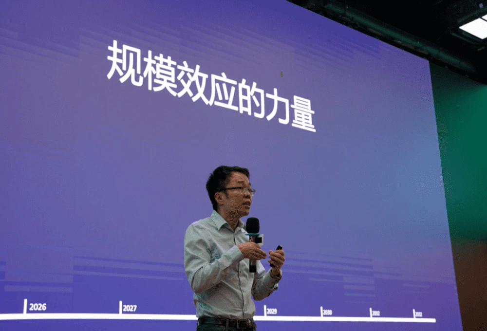 餐饮行业专家在TOA峰会发表"规模效应的力量"主题演讲：身着浅色衬衫的演讲者手持麦克风，身后紫色背景大屏幕醒目展示白色主题文字"规模效应的力量"，屏幕底部排列着2026-2030及2012等关键年份标识。画面采用中心构图突出演讲者，紫色背景与白色文字形成强烈视觉对比，配合舞台灯光效果，完整呈现中餐连锁化进程中规模经济效应的专业分析场景。屏幕年份序列暗示餐饮企业从单店经营（2012）到规模化发展（2026-2030）的成长路径，呼应文中MenuSifu提出的"六边形战士模型"中的规模化管理维度。