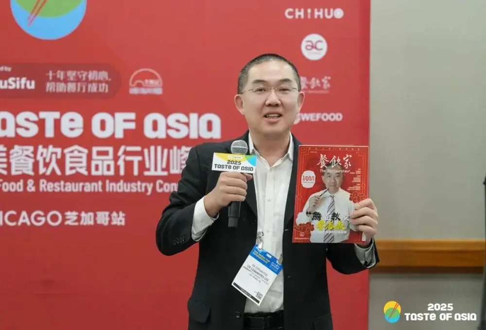 演讲嘉宾手持麦克风，拿着餐饮家杂志，在“2025 Taste of Asia”峰会芝加哥站演讲，背景为带有会议标识的红色展板，场地为会议厅。