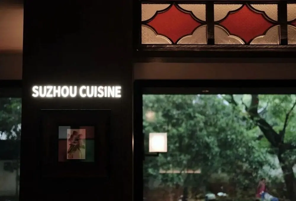 江南雅厨餐厅一角，墙上挂有“SUZHOU CUISINE”霓虹字样，窗外绿意盎然，呈现江南风情。