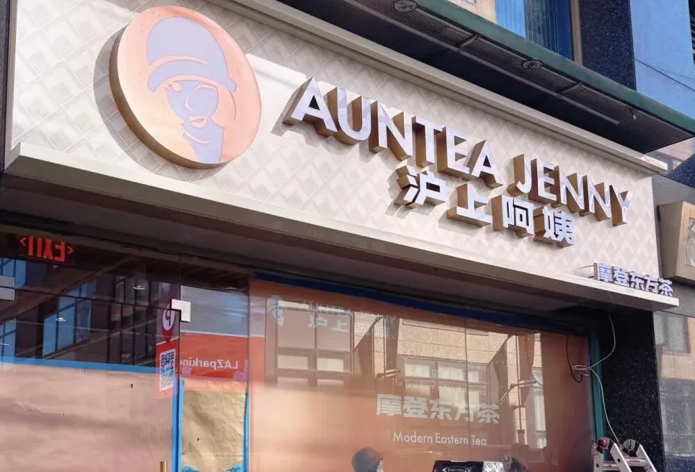 沪上阿姨纽约法拉盛门店装修现场，外墙上品牌中英文标识"AUNTEA JENNY 沪上阿姨"与"Modern Eastern Tea（摩登东方茶）"清晰可见，橱窗贴满"NYC"字样装饰贴纸，脚手架旁堆放着绿色梯子和银色建材。玻璃幕墙反射出纽约街景，与品牌东方美学风格的浅色招牌形成碰撞，呼应文中"美东首店即将落地"的全球化战略。