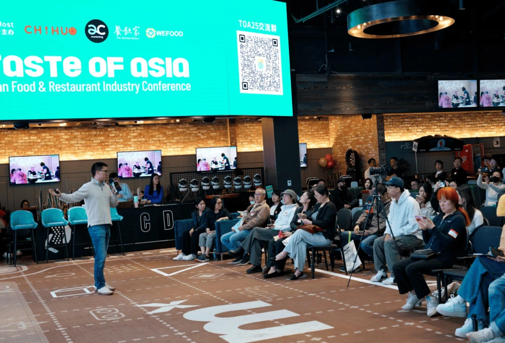 2025 TASTE OF ASIA餐饮峰会现场实况：一位演讲者手持话筒面向观众，背景大屏幕显示"TASTE OF ASIA"会议主题及主办方MenuSifu（WEFOOD）标识，右侧墙面悬挂实时转播显示屏。工业风砖墙搭配现代几何地砖，观众席坐满专注聆听的餐饮从业者，多人正使用手机记录精彩内容。屏幕细节显示"TOA25交流群"二维码及"Food & Restaurant Industry Conference"副标题，完整呈现这场汇聚中美餐饮精英的专业盛会，展现中餐产业资本化转型的前沿讨论场景。