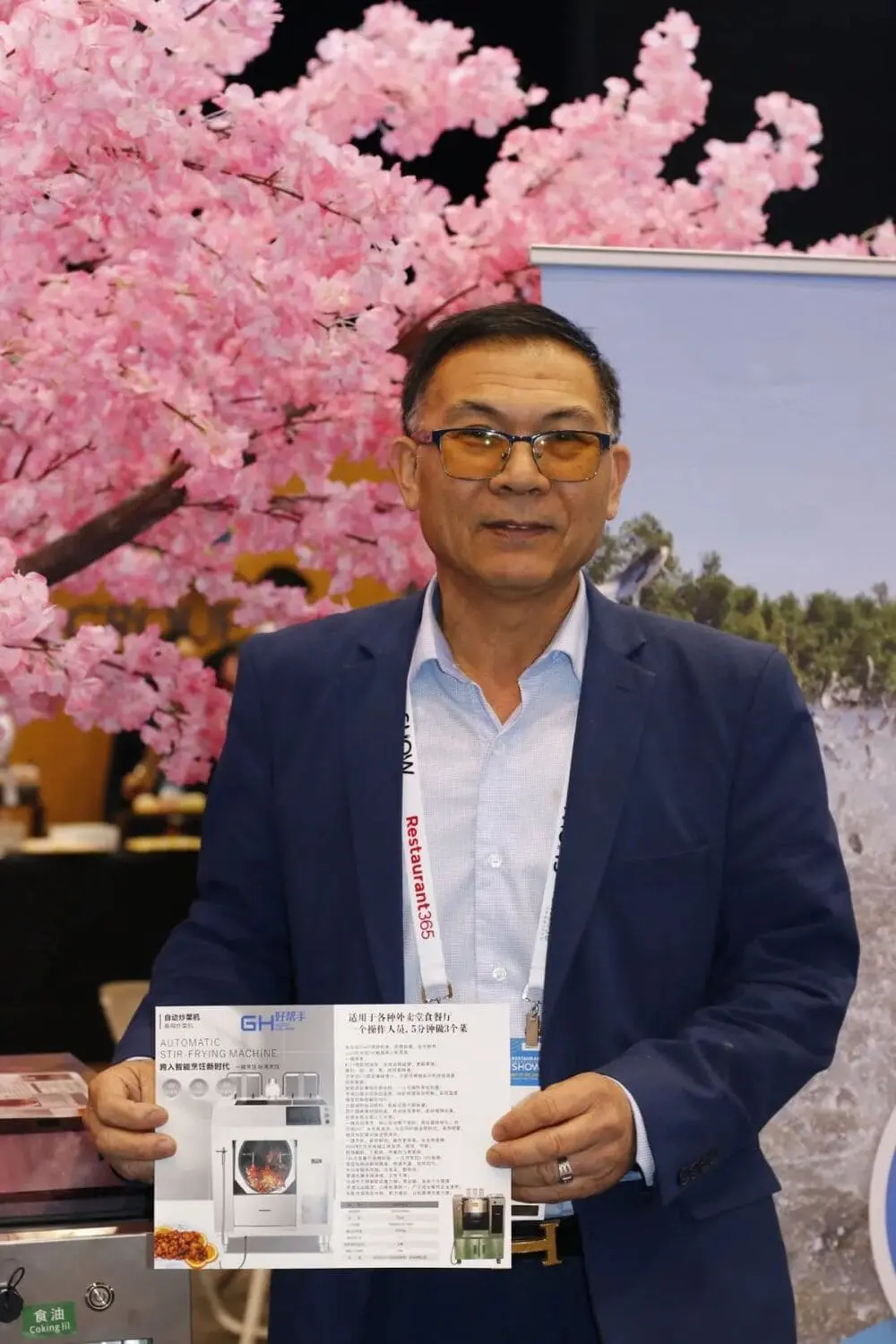 美中餐饮协会会长郑时坦向餐饮家展示“好帮手自动炒菜机“产品介绍彩页，背后是广告牌和樱花树