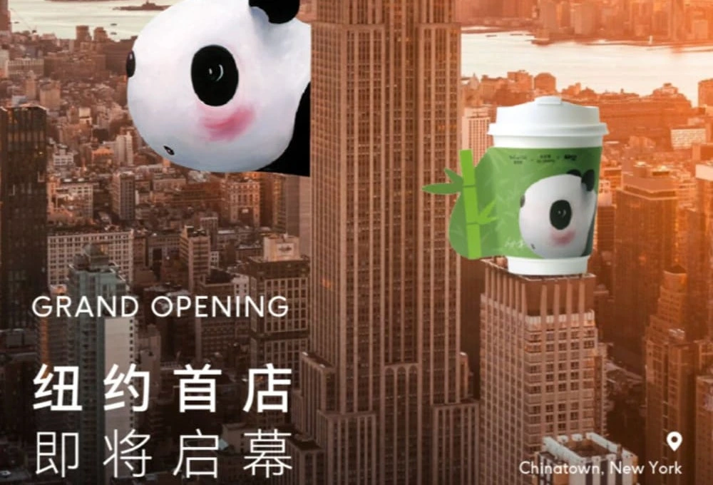 百分茶纽约首店开业海报：帝国大厦前，熊猫玩偶手持饮品杯，与品牌Logo“BeFine Cha”相映成趣。背景为黄昏下的纽约城市天际线，暖橙色调渲染开业氛围。海报标注“GRAND OPENING 纽约首店即将启幕”及地址“Chinatown, New York”，融合国潮元素与城市地标，传递活力与东方茶饮文化魅力。