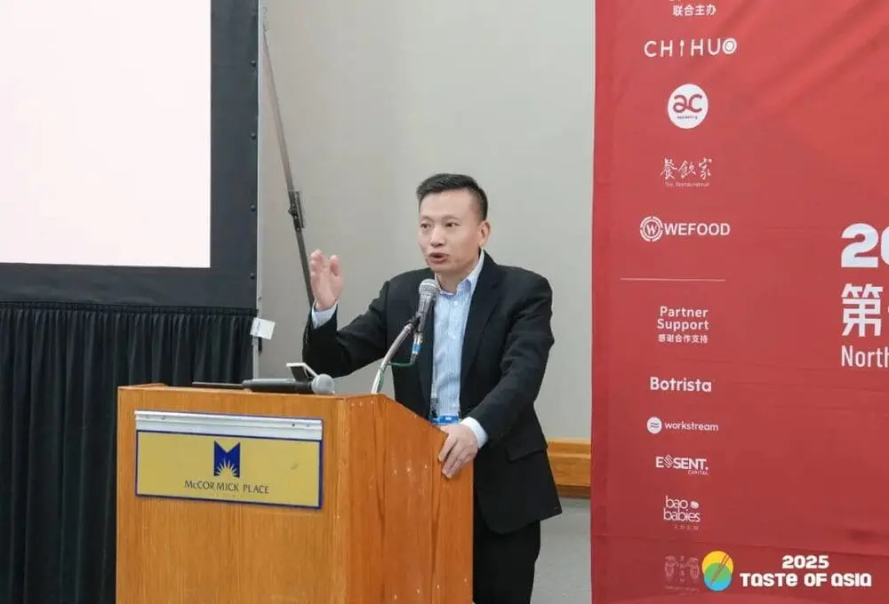 演讲嘉宾面对麦克风，在“2025 Taste of Asia”峰会芝加哥站演讲，背景为带有会议标识的红色展板，场地为会议厅。
