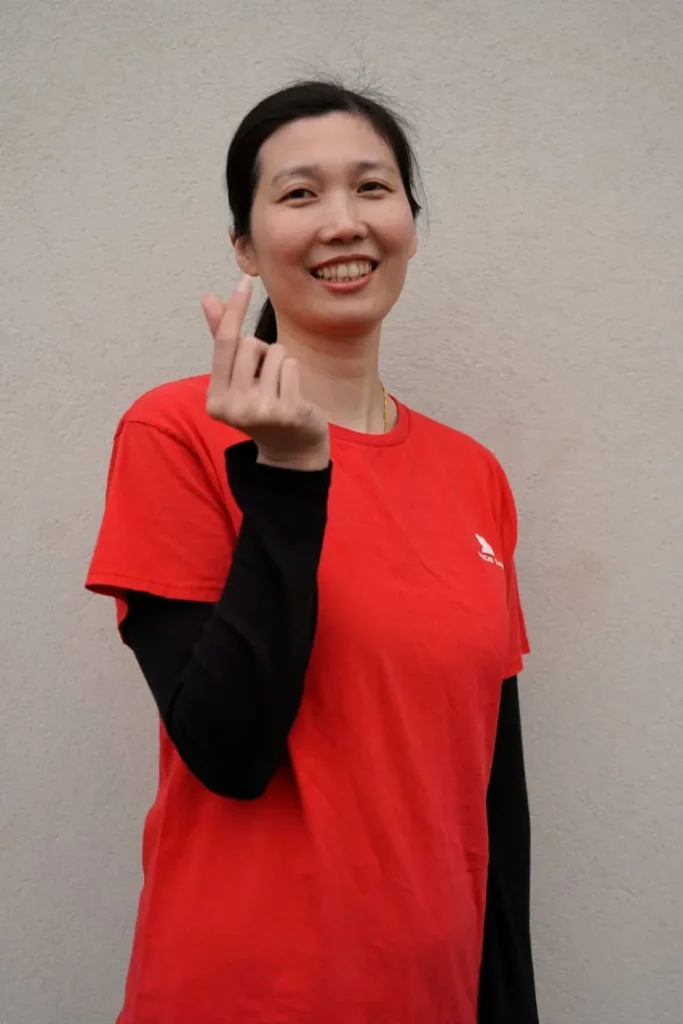 一位身穿红色 T 恤的女性面对镜头微笑，并用手指比出“爱心”手势，背景为浅色墙面。