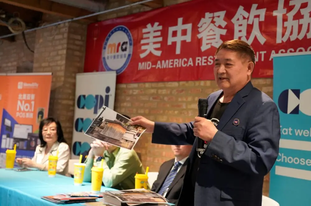 钟富华身穿深色西装，手持《图说自贡味道》图书对台下观众讲解，桌上摆放多杯奶茶和宣传资料，背景为美中餐饮协会展板。