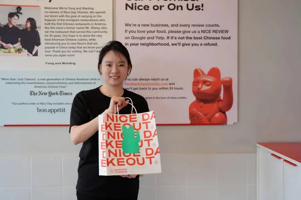 一位女性手持印有“NICE DAY TAKEOUT”字样的外卖袋，站在Nice Day Chinese餐厅的宣传墙前，背景有关于品牌创始人的介绍与顾客评价提示。