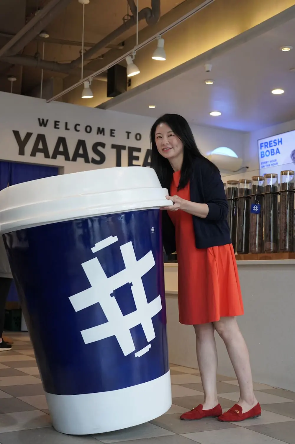 橙红裙装女士扶蓝白巨型奶茶杯模型，杯身印#字符号。背景为YAAAS TEA店铺，灯箱显示“FRESH BOBA”标语，透明料罐陈列柜清晰可见。