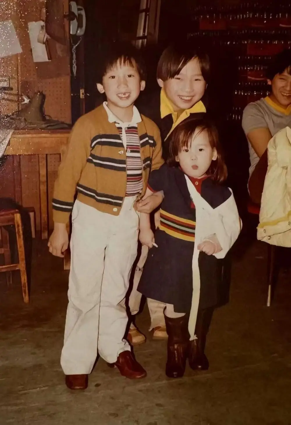 复古家庭照：童年陈国材（中穿黄领衣）、兄弟（左条纹开衫）与妹妹（右拼色外套）站于木质桌椅前，背景有厨房杂物，三人笑容纯真。