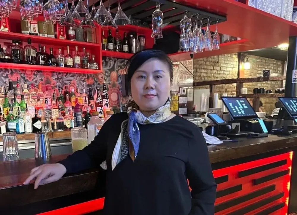 女士身穿黑色上衣和围巾，背景是餐厅就餐环境和酒柜，显得温暖雅致。