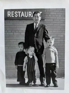 黑白老照片：陈国材祖父（西装）立于标有“RESTAURANT”的砖墙前，双手搭肩三个孙辈（Curtis、Chris和Craig），展现家族餐馆传承。