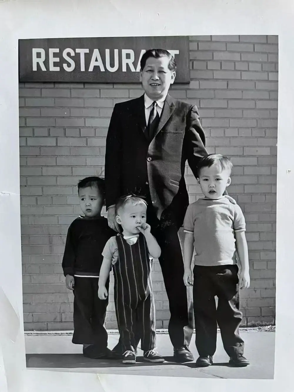 黑白老照片：陈国材祖父（西装）立于标有“RESTAURANT”的砖墙前，双手搭肩三个孙辈（Curtis、Chris和Craig），展现家族餐馆传承。