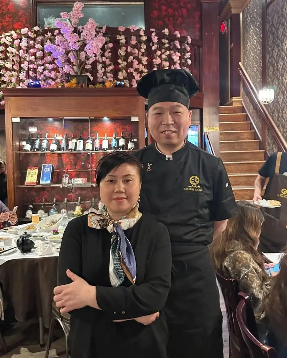 照片中是一位主厨和一位女士站在餐厅前厅，背后是摆放酒瓶的红色背景墙和粉色樱花装饰。