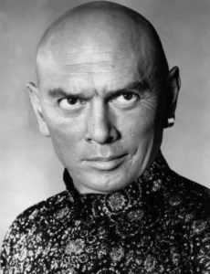 光头男星尤尔·伯连纳（Yul Brynner）黑白肖像：戴耳饰，穿繁复花纹高领衫，面容平静，浅灰背景突显其标志性形象。