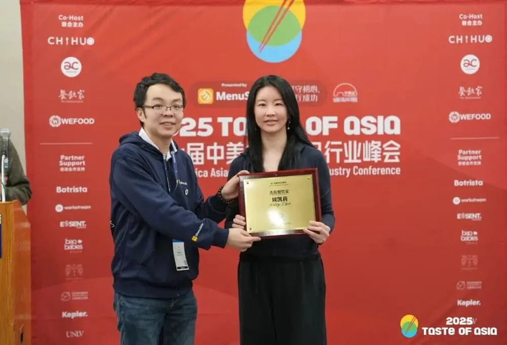 2025 Taste of Asia峰会现场，MenuSifu联合创始人李宇（左）向周晓婷颁发金色奖牌。她微笑注视刻有"杰出创业者"的牌匾，身后红色背景板印满赞助商Logo（含Botrista/CHIHU/WEFOOD），象征华人餐饮新生代突破。