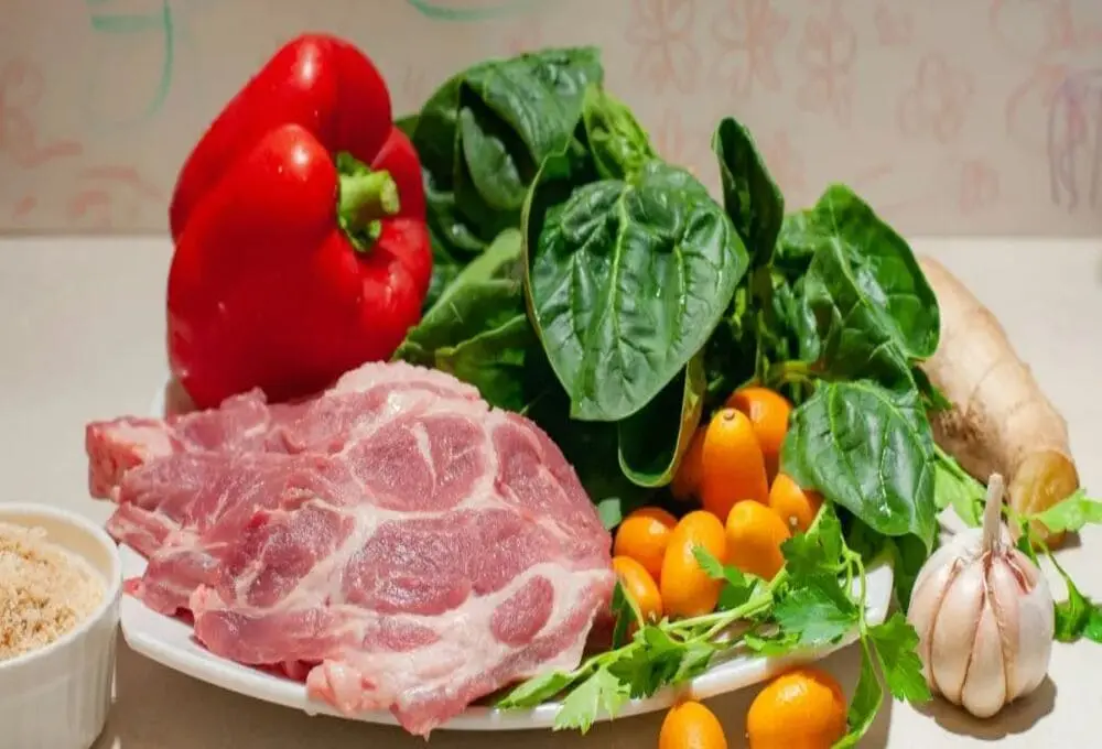 白色餐盘中红润猪肉片、鲜红甜椒、翠绿菠菜、嫩黄橘子瓣与姜蒜分区摆放，配调料碗。展示食材分类存储规范，符合食品安全标准中生熟分离要求，杜绝交叉污染风险。