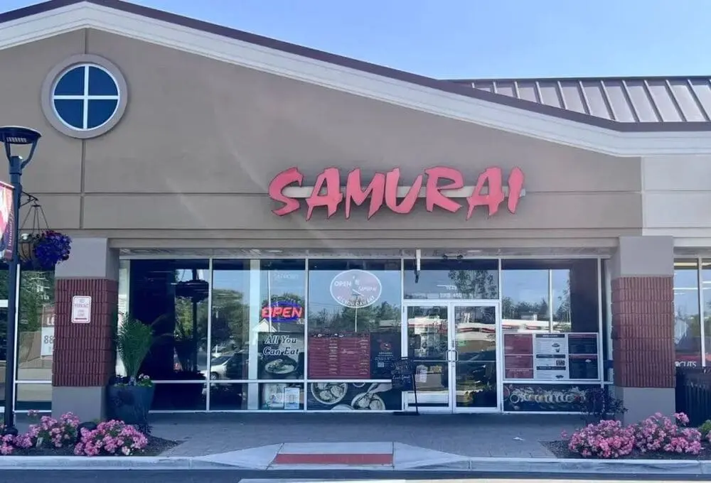 "Samurai Buffalo外立面：米色墙面配立体红字店招，落地窗贴有ALL YOU CAN EAT自助餐标识。门前粉色绣球花与石灯柱点缀，夜间OPEN灯箱亮起，彰显时尚日式自助餐厅定位。"