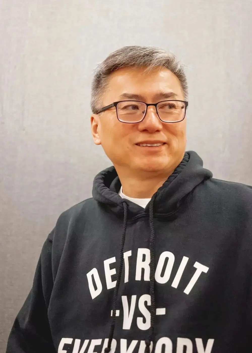 作家陈国材身穿印有“DETROIT VS EVERYBODY”字样的黑色连帽衫，侧身站立于灰墙前，灰白头发配戴黑框眼镜，神情专注。