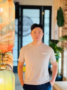 男性模特身穿浅米色Hurley短袖T恤，搭配深蓝长裤自然站立。左胸品牌logo清晰可见，背景含彩色灯笼、绿植及砖墙玻璃门，营造悠闲生活场景。服装剪裁简约，体现休闲时尚风格。
