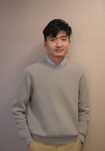 亚裔经营者双手插兜凝视远方，浅灰毛衣叠搭衬衫体现专业感，背景暖棕墙面暗示餐厅环境。代表出海品牌决策者正权衡供应链成本（美国厨师$6000月薪占售价60% VS 国内¥8000）与本土化策略（芝士虾仁饺改良），需用机械臂技术破局人工困局。