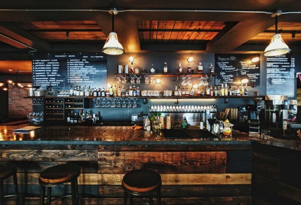 收购餐饮店需重点考察吧台设备：深色台面搭配木质酒架陈列约30个酒瓶及玻璃杯，两侧黑板清晰标注酒水菜单（含Draught Beer、Cider等12款价格），暖光吊灯与砖墙营造复古氛围。买家需核实酒类经营许可是否包含在转让范围内，并检查设备折旧状况。