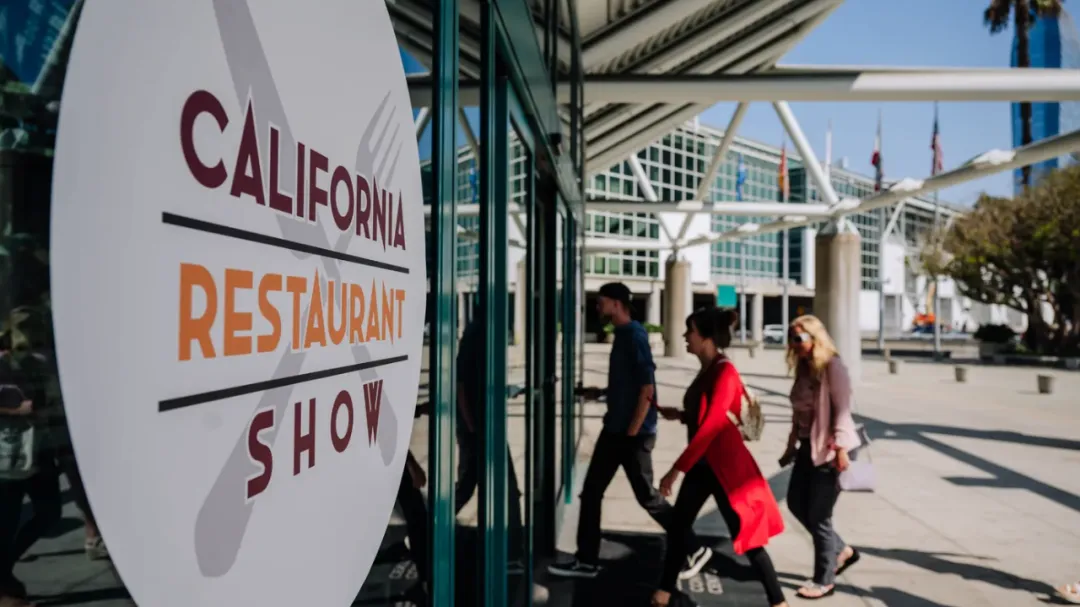 现代风格展馆入口特写：左侧玻璃幕墙镶嵌白色圆形「CALIFORNIA RESTAURANT SHOW」立体标志，透景可见人群步入建筑内部。建筑以大面积玻璃窗与白色钢构组成，顶部蓝天衬托60000平方英尺专业展馆空间。象征北美餐饮枢纽的门户价值，TOA亚洲专区入驻此地标性场馆。