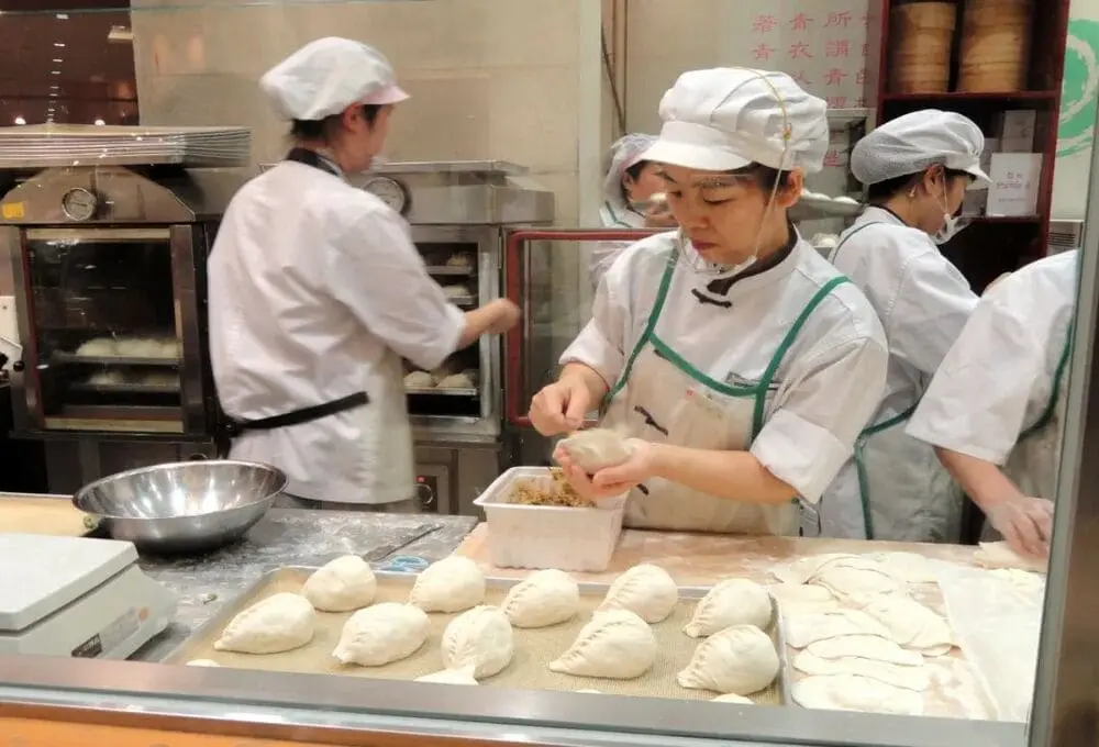 餐饮出海用工困境配图：厨房内三位厨师专注包制中式面点，近景人物正将馅料填入面皮，操作台摆放成排待煮的饺子与馄饨，蒸汽氤氲中展现人工密集型工序。身着标准白色工服与厨帽的团队分工协作，呼应文中高技术人力稀缺、工序流程化难等核心议题。