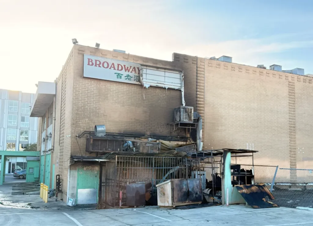 洛杉矶华埠"BROADWAY 百老汇"建筑火灾现场：一层入口金属结构焦黑变形，铁皮围栏倾圮，散落烧灼杂物。上层砖墙保留土黄原色，招牌边缘缺损。背景浅色现代高楼与晴空对比，记录2024年修复前实况，象征老店浴火重生历程。