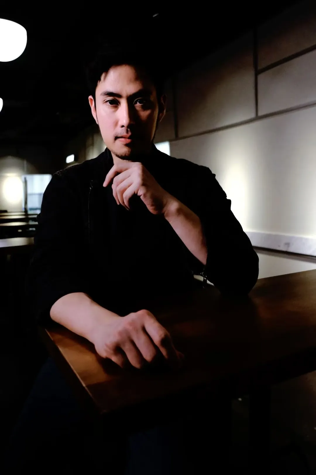 主厨Kenji穿厨师服单手托腮凝视镜头，黑背景突显轮廓。左颊光影分割面庞，右后方虚化料理台放置调料罐，象征传统技法与现代创新的沉思时刻。突出"外婆启蒙→纽约修业"的职业基因。